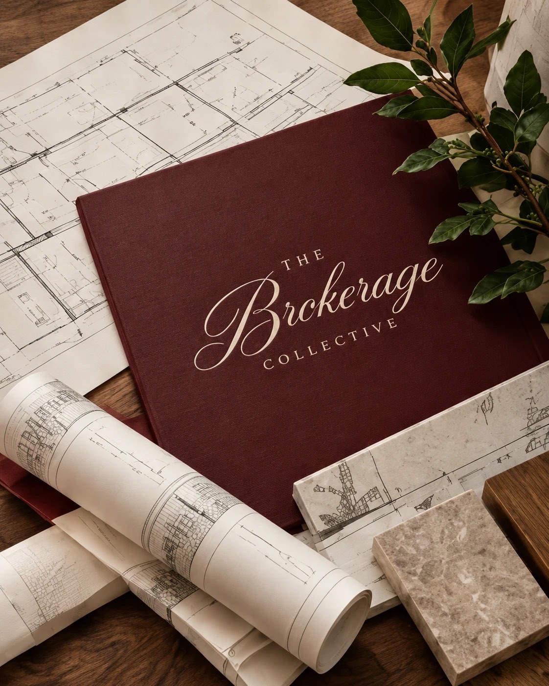A burgundy Brokerage Collective folio resting on house plans, with rolled drawings and stone samples
