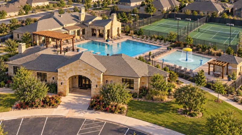 Aerial view of a stone clubhouse with a pool, splash pad and tennis courts, surrounded by homes